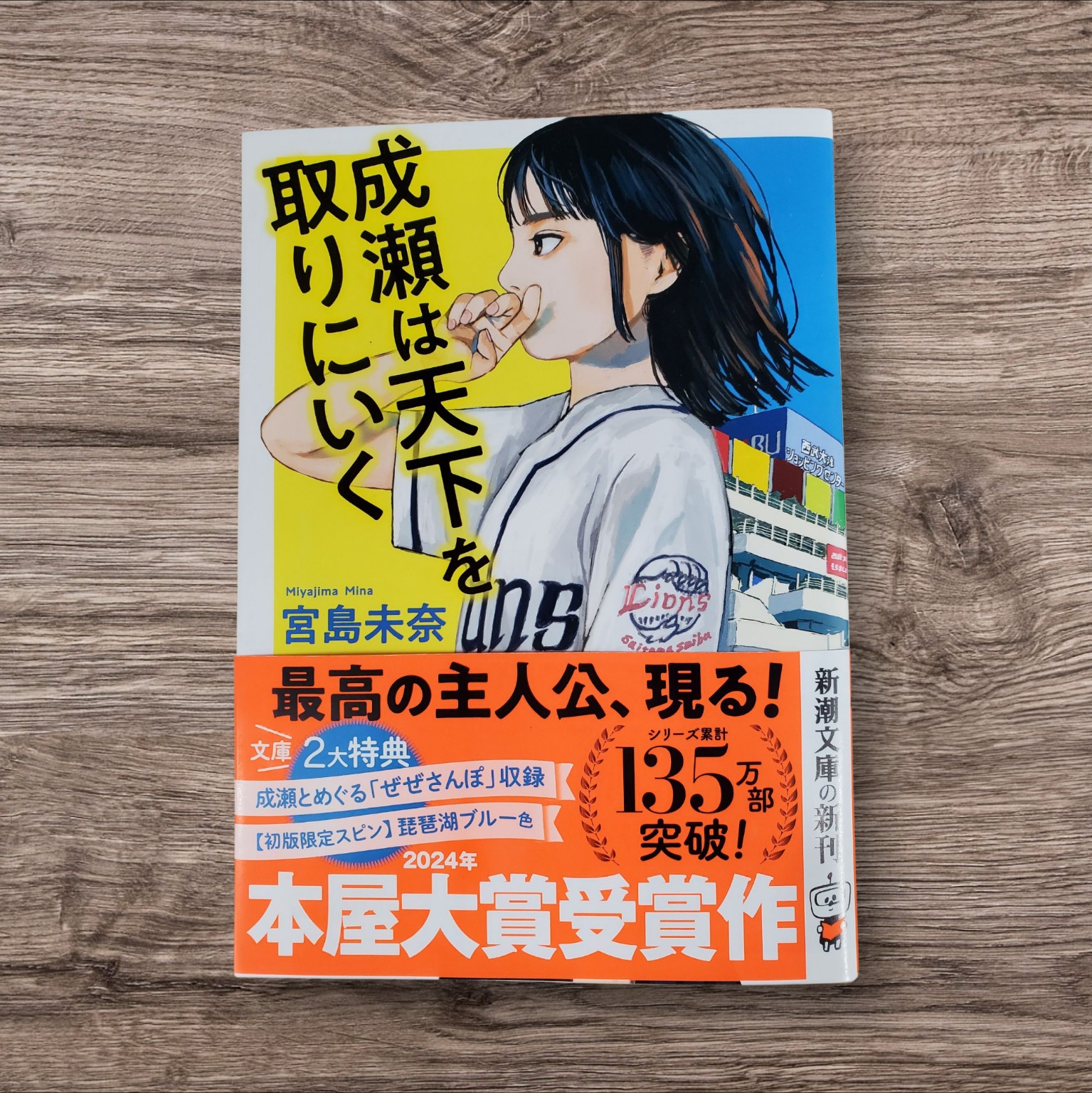 宮島未奈『成瀬は天下を取りに行く』文庫版の表紙。ユニフォーム姿で指笛を鳴らす成瀬あかりのイラストと、観光船ミシガンが描かれた背景が、物語の舞台である滋賀県大津の空気感を伝えるビジュアル。